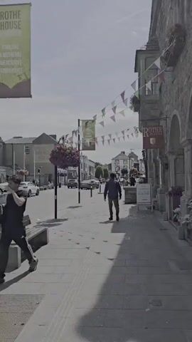 A man walks down a pedestrianized street lined with shops and buildings. He is the sole subject in the foreground, walking away from the camera. The street is paved with stone tiles, and a bench is visible on the left. Colorful triangular flags are strung across the street overhead, and banners hang from poles. 

On the right side of the street, a building has arched entrances and windows. A sign reads "ROTHE HOUSE" with an emblem. Another sign on a stand in front of the building says "EXHIBITION" and "The Restoration."

In the background, more buildings and parked cars are visible. Trees and flower baskets add decorative elements to the scene. The sky is cloudy but bright, suggesting it is daytime. The overall atmosphere appears calm and pleasant, typical of a town or city street on a fair-weather day.

Visible text:
- ROTHE HOUSE
- EXHIBITION
- The Restoration