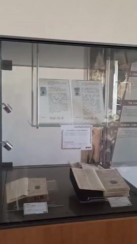 The video pans across a museum exhibition. In the foreground, there are glass display cases containing old books and documents. Behind the cases, large informational posters are mounted on the wall. One poster features a portrait of a man with a beard and a hat, accompanied by text in Italian. The room appears to be a hallway or common area with tiled floors and white walls. There is a doorway leading to another room. Further back, another glass display case sits on a table. Large windows let in natural light, illuminating the space. The overall impression is that of a historical or cultural exhibit.