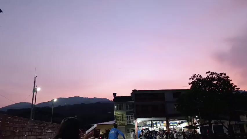 The video shows a tranquil evening scene with a sky painted in hues of pink and purple during sunset. The foreground features a street view with a few buildings and trees silhouetted against the vibrant sky. Distant mountains are visible on the horizon, adding depth to the landscape. Some people are present, though they appear as indistinct figures in the shadows. Streetlights are illuminated, casting a soft glow on the surroundings. The scene captures a peaceful moment, highlighting the natural beauty of the sky as dusk settles in.