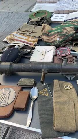 The video shows a table covered with various items, likely for sale at an outdoor market or flea market. In the foreground, there are several military-style helmets, including a blue one, a camouflage-covered one, and a khaki one. There are also military hats, a spoon, a small wooden box, and what appears to be a grenade launcher or similar large weapon. Behind these items, there are stacks of coins in protective cases, some books, a canteen, and bags. Further back, more tables are visible with more items, including what looks like a vintage arcade game or display, boxes, and chairs. The overall scene suggests a collection of vintage or historical items displayed outdoors.
