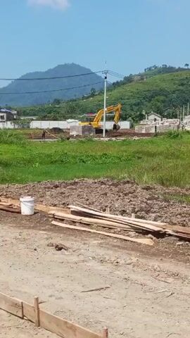 The video shows a construction site on a sunny day. An excavator is working in the distance, moving dirt. A person walks in the same direction. There's a pile of dirt, some wooden planks, and a white bucket in the foreground. In the very foreground there is also a wooden frame made from planks. Behind the site, a mountain is visible. Someone walks by the camera with a large bundle of green leafy vegetables in a cart.
