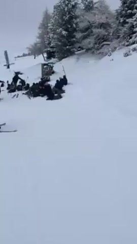 The video depicts a snowy ski area under a cloudy sky. A group of people, bundled in winter clothing, are gathered at the side of a ski slope, some sitting and others standing. Ski equipment, including skis and poles, is scattered around the group. The camera focuses on a person standing on skis, wearing a helmet, a gray hoodie, and green pants, holding ski poles. The camera then pans upwards to show the ski slope, bordered by orange safety netting, leading up to a line of snow-covered trees and a landscape of clouds in the distance. Other skiers are visible further up the slope.