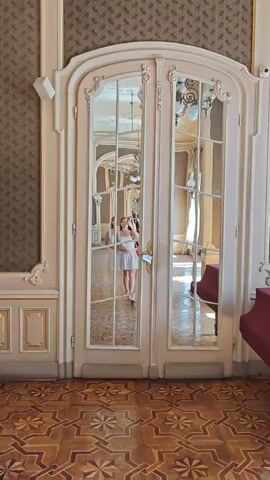 A young woman in a white dress with a polka dot pattern stands in front of double glass doors with decorative molding. She is taking a selfie in the reflection of the doors. Her hair is pulled back, and she is wearing sandals. The doors are paneled with glass and wood. Through the glass, a richly decorated room with ornate ceilings, chandeliers, and a red piano is visible. The floor is made of polished wood with a geometric pattern. The woman appears to be in a museum or a historical building.