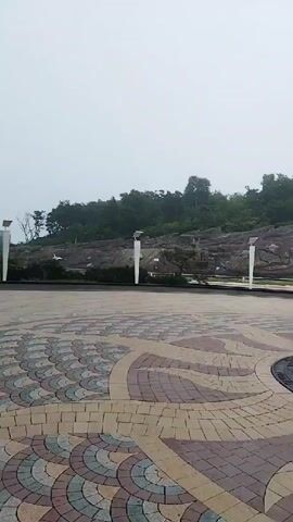 A person wearing a hat and blue jeans walks across a patterned plaza. In the background, there is a hill covered with trees. A sign is visible on the hill, and there are several lampposts on the plaza. The sky is overcast and foggy.