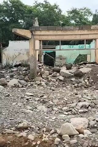 The video shows the aftermath of a disaster, likely a flood or landslide. A damaged building with a partially collapsed roof and walls stands amidst a large pile of rocks and debris. The building appears to be made of concrete blocks and corrugated metal. Trees and vegetation are visible in the background. The ground is covered with rocks, mud, and rubble. The sky is overcast. No people are visible in the video.