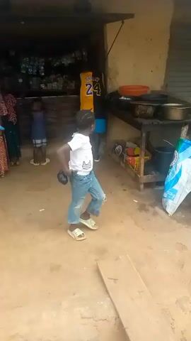 The video shows a young girl with braided hair wearing a white t-shirt and ripped blue jeans, walking across a rough, sandy ground. She is wearing light-colored slip-on sandals. In her right hand, she is holding a dark, cylindrical object. She appears to be walking towards the right side of the frame, occasionally glancing back.

In the background, there are several other people visible. To the left, a person wearing a yellow and black basketball jersey with the number "24" and the name "BRYANT" is partially visible. Further left, another young child in a blue shirt and shorts stands near a structure with shelves stocked with items. To the right of the main subject, there is a rustic wooden stall with various items on display, including what appear to be cooking pots, bags, and a broom. A large white and blue patterned bag is propped against the stall.

The setting appears to be an outdoor marketplace or a busy street area in a village or town, given the unpaved ground, simple structures, and open-air stalls. The lighting suggests it is daytime, possibly late morning or afternoon, with no clear indication of specific weather conditions. The overall atmosphere seems casual and everyday. There are no discernible emotions conveyed by the main subject or other individuals.

Visible text includes "airtel" on a sign in the background, suggesting a telecommunications provider. The scene is set in Madakiya, Nigeria.