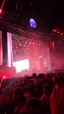 The video shows a crowded outdoor concert with a large stage and multiple video screens displaying vibrant graphics. A DJ is visible on stage, silhouetted by bright lights and smoke. The audience is enthusiastically engaged, with many people raising their hands in the air. The stage features a large, circular logo at the top. The lighting is dynamic, with red and white hues dominating the scene. The overall atmosphere is energetic and festive.