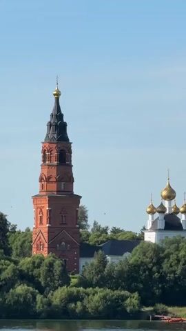 The video captures a scenic view of a monastery with a tall, red brick bell tower and a white church with golden domes, set against a clear blue sky. The buildings are surrounded by lush green trees and foliage. In the foreground, a body of water, likely a river, is visible, with a small portion of a wooden dock and a red boat on the right side. The overall impression is one of tranquility and historical significance.