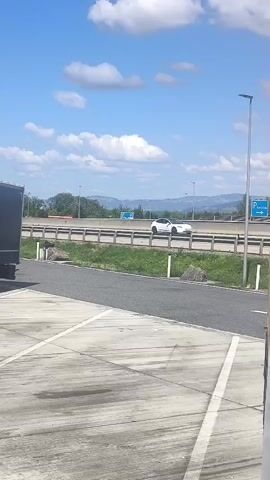A truck pulls into a parking lot. The truck has a black cab and a silver trailer. It is a sunny day with blue skies and white clouds. There is a highway in the background with cars and trucks driving on it. There are also trees and mountains in the distance. The parking lot is made of concrete and has white lines painted on it.