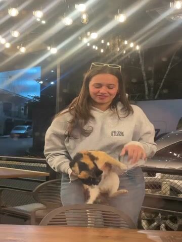 A young woman is outdoors at night, on a patio that is lit with strings of light bulbs. She is wearing a light grey sweatshirt, blue jeans, and has sunglasses perched on top of her head. She is holding a calico cat. She bends over, looking at the camera, and smiles. She then releases the cat, which jumps off of a chair. The woman laughs.