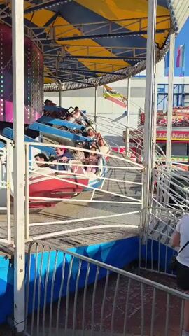 A crowd of young people is enjoying a fairground ride. The ride appears to be a spinning amusement ride with multiple cars that tilt. The people in the cars are laughing and smiling, enjoying the thrill of the ride. The ride is decorated with colorful lights and has a large, tent-like canopy overhead. The background shows a typical fairground setting with other rides and stalls visible. The sun is shining, indicating it is daytime.