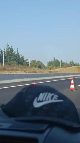 A first-person perspective video taken from inside a vehicle shows a highway with traffic cones marking a lane closure. The camera is focused on a black Nike baseball cap resting on the dashboard in the foreground. In the background, the road stretches out, with several orange and white traffic cones placed at intervals. A gray car is visible ahead, driving in the same direction. The surrounding environment consists of a clear blue sky, sparse vegetation, and distant trees.