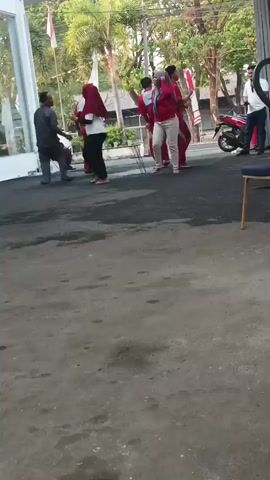 This is a video of a group of people. There are five individuals visible. One person is wearing a grey shirt and dark pants. Another person is wearing a red shirt with white accents and white pants. This person appears to be female and is wearing a headscarf. Two other individuals are wearing similar red outfits. One individual is wearing a white shirt and dark pants. The group appears to be interacting in an outdoor setting. There is a sidewalk or pavement area with a rough texture. In the background, there are trees, buildings, a truck, and a motorcycle. One of the individuals in red is holding a small object, possibly a microphone, and appears to be singing or speaking. The others are observing or participating in some activity. The overall mood seems lighthearted and energetic.