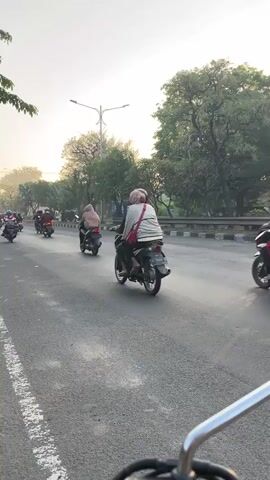 The video shows a street scene with a considerable number of motorcycles and scooters traveling in the same direction. The road appears to be paved with asphalt and has a white dashed line marking the lane. There are trees and foliage lining the side of the road, and in the background, there is a hazy sky, suggesting it might be early morning or late afternoon with some atmospheric conditions. Several individuals are riding the motorcycles, some wearing helmets and others not. One person in the foreground on the left is riding a black motorcycle and is wearing a cap and shorts. In the middle of the frame, several other motorcycles are visible, with riders wearing various clothing, including a person in a pink shirt and another in a blue jacket. On the right side of the road, more motorcycles are also seen. The overall impression is of a busy urban or suburban road with a significant volume of motorcycle traffic.