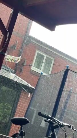 The video shows a heavy rain shower from the shelter of a wooden porch or pergola. The camera is pointed outwards towards a red brick house, a trampoline, and a bicycle. Rain is clearly visible falling heavily across the entire scene.  A washing line strung with clothes and pegs is attached to the side of the house. The trampoline, fitted with a black safety net, stands in the foreground.  A bicycle is parked to the right, partially obscuring the view. The scene appears to be a backyard during a downpour.