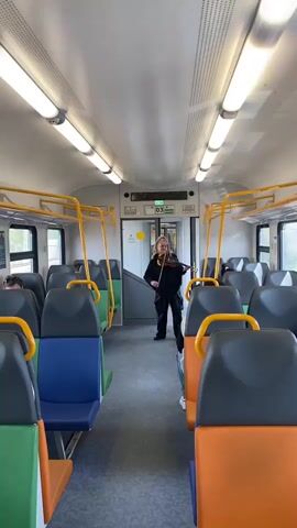 The video shows a woman playing the violin in the aisle of a train. The woman is dressed in black and stands in the center of the train car, with rows of mostly unoccupied seats on either side. The seats are varied in color (blue, orange, green). Yellow poles support the overhead compartments. The train interior is well-lit by overhead fluorescent lights. Some passengers are visible in the background. The destination indicator above the door is in green.