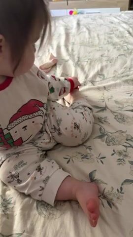 A small child in white pajamas with a holiday-themed cartoon character print sits on a bed covered with a patterned floral-print comforter. The child looks towards the side before turning to look directly at the camera. The child has fair skin and brown hair, and a small white clip in their hair. Several other small objects can be seen on a table or surface in the background, blurred.