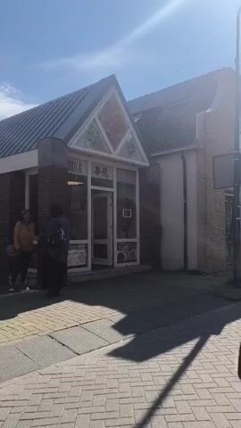 The video shows a street scene on a sunny day. On the left, there is a building with an entrance to a Chinese restaurant. The restaurant has a sign with Chinese characters and the word "CHINA". Two people are visible near the entrance, one standing and the other walking away. The street is paved with cobblestones. To the right of the restaurant, there is a two-story brick building. Further down the street, there are other buildings with shop fronts. The camera appears to be moving, possibly from a vehicle, as indicated by the reflection in what looks like a side mirror at the bottom right of the frame. The sky is blue with some clouds. The overall impression is of a quiet street in a town or village.