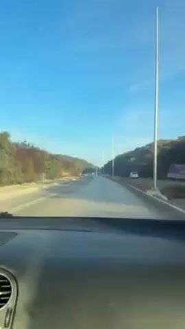 The view from inside a car driving on a road. The sky is blue, and there are trees on either side of the road. There are tall light posts along the roadside. A white car is parked on the side of the road. There are road construction signs on the side of the road.