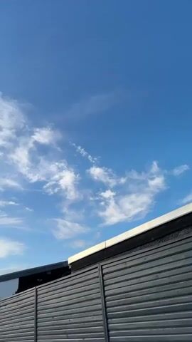 The video shows a view of the sky and buildings from street level. The sky is blue with scattered white clouds that appear wispy and feathery. In the foreground and midground, there are residential buildings with peaked roofs. One building has a dark metal fence or gate in front of it. Another building has a white facade with a visible house number "19". There are also cars parked along the street. In the distance, a tall, dark steeple of a church is visible. The sun appears to be low in the sky, casting shadows and illuminating the clouds. The overall impression is that of a quiet, somewhat overcast day in a European town.