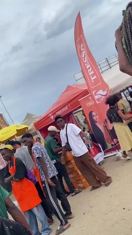 The video shows a gathering of people outdoors, likely at a market or festival. Several individuals are visible, some wearing aprons, suggesting they may be vendors or staff. One person is carrying a large bag on their head. There are tents and umbrellas set up, providing shade. Banners with the word "TRESSA" are displayed. The sky is overcast. The general atmosphere appears lively and crowded. The camera is moving.