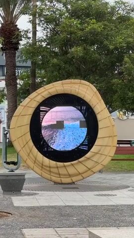 A video captures a large, circular digital display framed by a textured, organic-shaped wooden structure. The display cycles through various scenic images, including a coastal town, a field of red flowers, and a graphic with text reading "ARONA." The setting appears to be an outdoor public space, with a palm tree on the left, lush green trees in the background, and a bench visible behind the display. The ground is paved with large stone tiles and gravel. A small black object, possibly a trash receptacle, is situated to the left of the display.