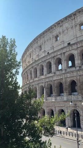 The video pans across the exterior of the Colosseum in Rome. The ancient amphitheater, made of stone, shows its characteristic arched openings on multiple levels. Trees with green leaves partially obscure the left side of the structure. In the background, a clear blue sky is visible. Below the Colosseum, a street runs alongside it, with a sidewalk and a few people walking. A lamppost stands near the sidewalk. The video is shot from a slightly elevated perspective.