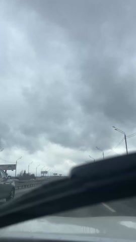 The video shows a view from inside a car driving on a highway on a cloudy day. The sky is overcast with thick, grey clouds. Streetlights line the sides of the highway. Other cars are visible on the road, and some trees can be seen in the distance. The windshield wipers are briefly activated to clear rain or road spray.