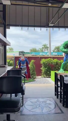 The video shows an outdoor seating area of a restaurant or cafe. In the foreground, there are several tables with chairs, some set with placemats and glasses. To the left, a large statue of Superman stands on a platform. To the right, a similarly sized statue of the Hulk is positioned. Beyond the statues, there is a red wall with some greenery and a fountain. In the background, there are electrical wires, a street, and some buildings. A few people can be seen in the background, some interacting with the statues or working. The overall atmosphere appears to be casual and outdoors.