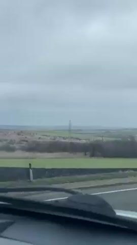 The video shows a view from inside a car driving along a road. Outside the car, the landscape is visible, consisting of fields of green grass, interspersed with patches of brown vegetation. In the distance, power lines and utility poles are visible. The sky is overcast and grey. The road itself is paved with a white line marking the lane. The car's dashboard and windshield wipers are partially visible in the foreground.