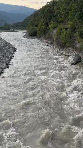 A wide, murky river flows through a valley with forested hills on the right bank and a rocky embankment on the left. The water is turbulent, with visible ripples and waves. The sky is overcast, suggesting either dawn or dusk. The scene is a natural landscape, devoid of any people or man-made structures apart from the possible retaining wall along the left bank.