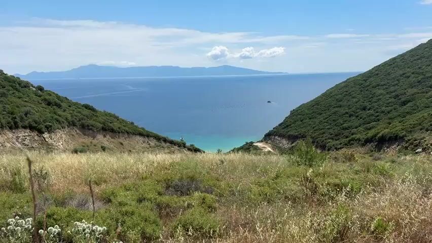 This video captures a scenic view of a body of water, likely the sea, bordered by lush green hills on either side. The foreground is dominated by dry, golden grasses and scattered green vegetation. The terrain slopes downwards, revealing a narrow path or road on the right side, and a more rugged, eroded landscape on the left.

In the distance, a vast expanse of deep blue water stretches out, meeting a hazy, mountainous island range on the horizon under a clear blue sky dotted with fluffy white clouds. The water closer to the shore transitions to a lighter, turquoise hue, suggesting shallow depths. A small white boat is visible near the shore, and further out in the water, a cluster of dark objects, possibly buoys or a small raft, can be seen. A single figure, appearing to be a person, stands near the turquoise water on the left side of the frame. The overall impression is one of a tranquil, natural landscape, possibly a coastal viewpoint.