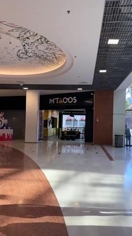 The video shows a commercial establishment with a sign that reads "M'TACOS AUTHENTIC FRENCH TACOS". The interior of the establishment features a counter with a menu board and seating area with sofas and small tables. In the foreground, two individuals are walking past the establishment. The environment appears to be a mall or a similar indoor public space with a tiled floor and ceiling lights.