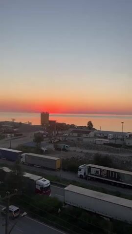 This is a vertical, outdoor video taken during a sunrise. The sun, a bright orange disc, is low on the horizon, casting a warm glow over the water and the sky. The sky transitions from a pale blue at the top to shades of orange and pink near the horizon. In the middle ground, there is a body of water, likely the sea, with a distant shoreline. Along the coast, there are industrial buildings, possibly a port or a construction site, with structures like silos and cranes visible. A highway runs parallel to the coast in the foreground, with several large trucks and a few cars driving on it. Some of the trucks are parked in a lot adjacent to the highway. The overall scene depicts a coastal industrial area at dawn, with the natural beauty of the sunrise contrasting with the man-made structures.