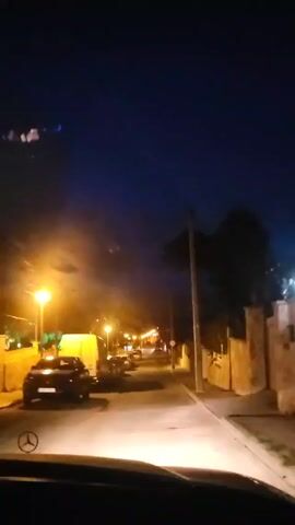 The video shows a nighttime street scene from the perspective of someone driving a car. There are parked cars on the left side of the street and a stone wall on the right. Streetlights illuminate the road. A speed limit sign of 30 is visible on a pole to the right. The sky is a deep blue.  A tree is visible on the right side of the street.  The car's headlights shine down the road. A Mercedes logo is visible on the steering wheel.  Music plays in the background.
