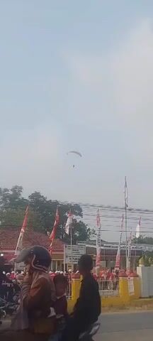 A paraglider is visible in the sky. Below, a building with a red roof is seen, adorned with Indonesian flags. A group of people are gathered in front of the building, and several vehicles, including cars and motorcycles, are present on the road. The sky is hazy and overcast.