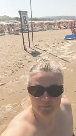 A woman with blonde hair pulled up and dark sunglasses is taking a selfie on a sandy beach. She is wearing a bikini top. The background shows a crowded beach with lounge chairs, umbrellas, and people swimming in the ocean under a clear blue sky. The camera pans slightly to the right, showing more of the beach scene with people walking along the shoreline and in the water.