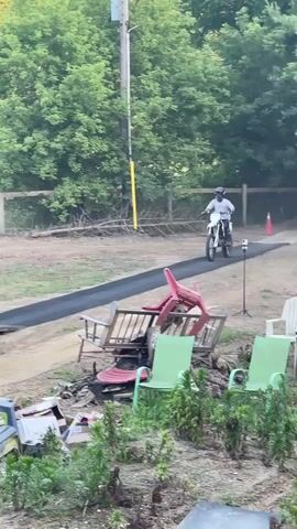 A person on a dirt bike rides up a ramp and launches into the air, performing a trick. The person then lands on an inflatable airbag designed for stunts. The video shows trees and a clear sky in the background. There are also some discarded chairs in the foreground.