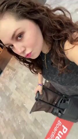 A young woman with long, curly brown hair and pink lipstick is shown in a selfie video. She is wearing a dark grey sleeveless top, a gold-colored necklace, and a purple bracelet on her wrist. She holds the handle of a black rolling suitcase, which has a brown bag resting on top. The setting appears to be an indoor space with a light-colored tiled floor. A red strip with white text, partially visible as "de Ab", and a white arrow marking a path is on the floor. She looks towards the camera, occasionally smiling, suggesting she is in a transit area, likely an airport in Apodaca, Mexico.