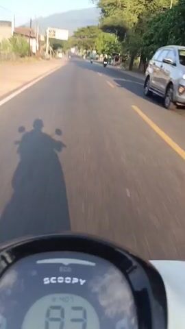 This is a point-of-view video taken from a scooter on a paved road in Sumberanyar, Indonesia. The sun is low in the sky, casting long shadows from the scooter and its rider onto the road. The rider's shadow is prominent in the foreground, showing the silhouette of a person on a scooter. The scooter's dashboard is visible in the bottom of the frame, displaying "SCOOPY" and a digital readout showing the speed as 83 km/h, then 82 km/h, and 84 km/h.

The road itself is asphalt with a yellow line marking the center. On the left side of the road, there are buildings, some appearing to be shops or houses, and vegetation. On the right side, there are more trees and some small structures. In the distance, a mountain range is visible under a clear, bright sky.

Several other vehicles, mostly scooters or motorcycles, can be seen on the road ahead, moving in the same direction. The overall scene suggests a rural or semi-urban environment during what appears to be either early morning or late afternoon, given the low angle of the sun and the warm light. The activity is a scooter ride along a road, likely a commute or a journey through the area. The mood appears relaxed and peaceful, with the rider enjoying the ride and the scenery. No discernible emotions are conveyed beyond the sense of movement and travel.