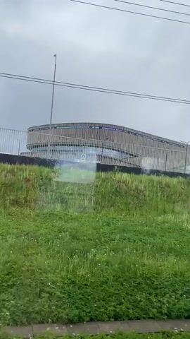 The camera captures a wide shot of a structure with a distinctive design, featuring a rounded facade constructed of vertical wooden slats. A railing surrounds the perimeter of the structure, suggesting it's elevated or on a raised platform. The sky overhead is overcast and gray. In the foreground, there's a lush, green field of vegetation, growing on what appears to be a hillside. Power lines are visible across the upper portion of the frame. The camera seems to be in motion, possibly from a vehicle passing by, indicated by the slight blurring of the grass and the changing perspective.