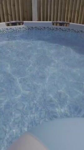 This media file shows a close-up view of an inflatable pool. The water is clear and blue, with sunlight reflecting off the surface, suggesting it is daytime and likely sunny weather. The pool has a decorative tiled edge and is surrounded by a wooden fence, which provides a backdrop. In the center of the pool, a multi-colored beach ball is floating. The ball has segments of white, yellow, pink, and light blue. The scene is peaceful and appears to be set in a backyard or garden area. There is no visible text within the media file.