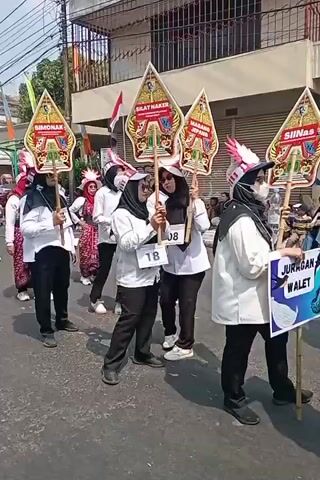 A group of people in a parade. They are wearing white shirts, black pants, and hijab. Some of them are holding up large, colorful flags that appear to be a traditional Indonesian art form called Wayang Kulit. There are other participants in the parade behind them, as well as spectators on the side of the road. The setting appears to be a street in a city.