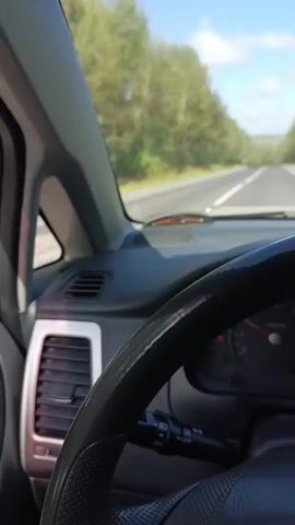 The video is filmed from the perspective of a driver in a car. The dashboard and steering wheel are visible in the foreground. The road stretches ahead, lined with trees on either side. The sky is blue with scattered clouds. The car appears to be moving along a paved road, possibly in a rural or suburban area, surrounded by a forest. The lighting suggests it is daytime.