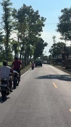 The video shows a road with two people on motorcycles. The person in the foreground is on a scooter, and the person behind them is on a motorcycle. Both are riding away from the camera. The road is paved and has a yellow line down the center. There are trees on both sides of the road, and some buildings in the distance. The sky is blue and clear.