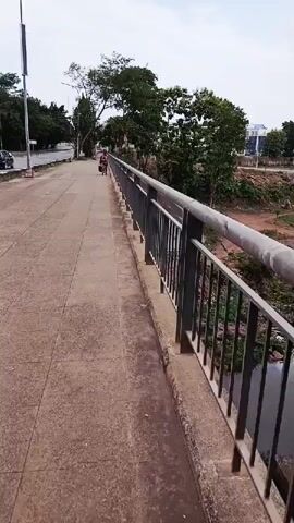 The video shows an outdoor scene, likely a bridge crossing over a body of water, in Kitwe, Zambia. The camera is positioned on a pedestrian sidewalk, looking forward along its length.

To the right, a metal railing, typical of a bridge, runs parallel to the sidewalk, separating it from the waterway below. The water itself is dark and mostly obscured by the railing. Beyond the railing, there's a sloped embankment with some sparse vegetation and reddish soil.

On the left side of the frame, a paved road for vehicles runs parallel to the sidewalk. A street light pole is visible near the beginning of the road. In the distance, a dark-colored car can be seen on the road.

Further down the sidewalk, a person is walking away from the camera. They are dressed in a patterned garment and carrying what appears to be a bag or basket.

Trees with green foliage are scattered along the left side of the road and also visible further in the background, suggesting a somewhat green environment. In the far distance, beyond the trees, some buildings with light-colored walls and dark roofs can be faintly seen under an overcast sky. The overall weather appears to be cloudy but bright, indicating daytime. No visible text is present in the video.