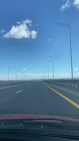 The video captures a forward-facing view from inside a moving vehicle on a multi-lane highway or bridge. The scene is dominated by a bright blue sky with scattered white cumulus clouds. A long, straight asphalt road stretches into the distance, marked with white and yellow lines, including a solid yellow line on the right and a broken white line on the left, with a thin red line also visible on the left side of the lane.

Numerous street light poles line both sides of the road, extending far into the horizon, creating a sense of depth and perspective. Guardrails are visible along the edges of the road. In the far distance, there appears to be a body of water or flat landscape under the open sky. Another vehicle, possibly a truck, is faintly visible in the distance on the highway. The dashboard of the car from which the video is filmed is partially visible at the bottom of the frame. The weather appears clear and sunny. The location is Soroch'i Gory, Russia.