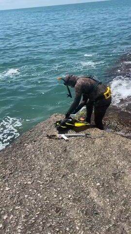 A person in a camouflage wetsuit, snorkel mask, and fins crouches on a rocky shore next to a body of water. The person is reaching down towards a pair of yellow fins and a black bag. A spearfishing gun with a white handle lies on the rocks in front of the person. The water is blue and appears to be choppy, with small waves breaking on the shore. The sky is clear and the sun is shining.