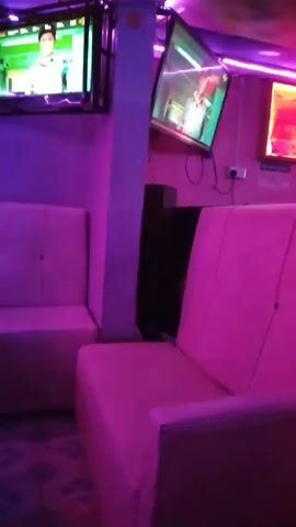 Here is a description of the video:

The video depicts the interior of a dimly lit, potentially private, space, possibly a bar or lounge, characterized by bright neon purple lighting. The main focus is a set of two white sofas arranged to create a small seating area. 

Several objects are visible in the room, including two televisions mounted on the wall, each displaying different content, though it is hard to ascertain the specific content on the screens. Additionally, there's a neon sign or display on the wall and a black speaker or component positioned behind one of the sofas.

During the video, the camera pans slightly, revealing two people sitting on the sofa furthest from the camera. Both appear to be casually dressed and engaged in conversation. A person wearing a blue hijab and carrying a pink bag walks between the camera and the group of seated men, obscuring the view. Later, the camera reframes the seating area again.
