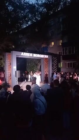 The video captures an outdoor nighttime event at what appears to be a community gathering. A stage is set up under an archway with illuminated text that reads "АЛЛЕЯ МОЛОДЕЖИ" (Alley of Youth). In front of the archway, a female performer in a light-colored outfit is singing into a microphone, with a speaker positioned to her left. A crowd of people, silhouetted against the stage lights and the building behind, are gathered to watch. Some individuals in the audience are holding up what appear to be lit candles. The environment is dark, with trees visible in the background and a multi-story building behind the stage, its windows illuminated. The performer gestures with her free hand as she sings, and the audience seems engaged with the performance.